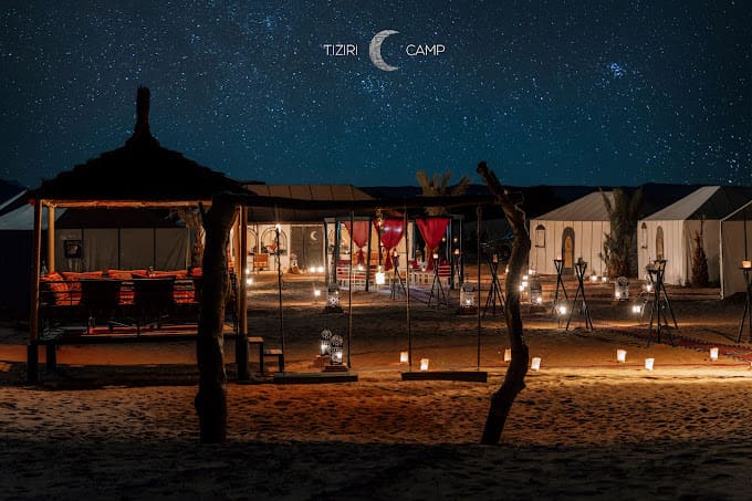 Desert camp at night under a clear starry sky with a crescent moon, lit tents and lanterns on the sand near Merzouga