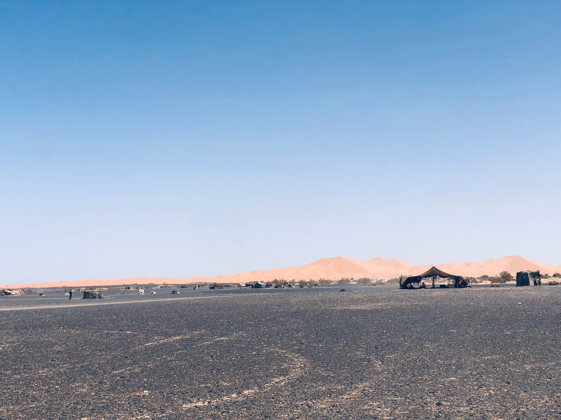 Flat, dark stony desert plain with a nomad tent, and the orange Erg Chebbi dunes rising far behind it